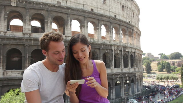 Couple using smart phone in Rome by Colosseum looking at pictures or using travel app in Italy. Happy lovers on honeymoon sightseeing Coliseum. Love and travel concept with multiracial couple.