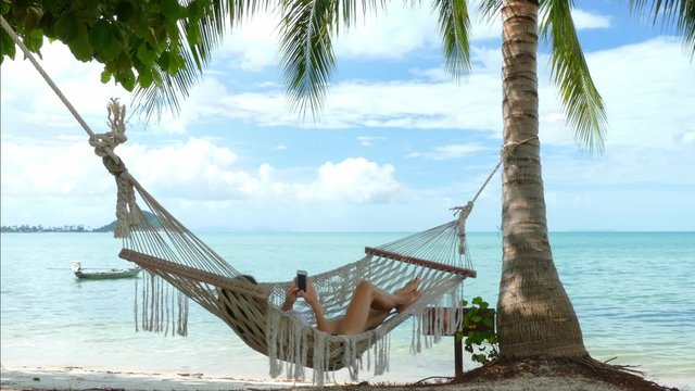 Woman using mobile phone in a hammock on the beach