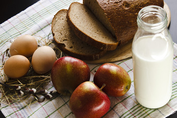 Country still life with food farming. Milk, bread, eggs, red apples and a sprig of willow on a towel on a black background. Easter still life.
