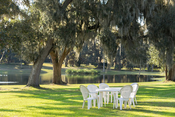 White plastic table and chairs outside in a garden on green lawn by a pond or lake in the afternoon sun and a peaceful relaxing serene tranquil setting - 103211017
