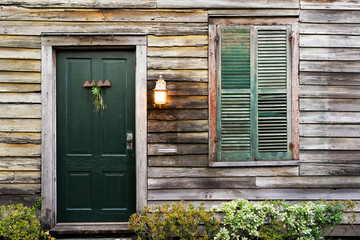 Old rustic vintage dilapidated antique house home building structure with green intricate front door and window with shutters closed and porch light lantern glowing turned on - 103213620
