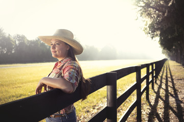 Cowgirl woman in cowboy hat flannel shirt and jeans leaning on country rural fence looking...