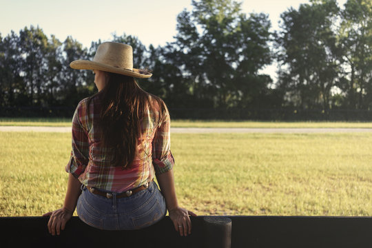 Cowgirl lady woman female wearing cowboy hat and flannel shirt with jeans sitting on country rural fence by a horse pasture paddock looking confident happy serene smart alone waiting watching patient