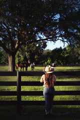 Cowgirl lady woman female wearing cowboy hat and flannel shirt with jeans leaning on country rural fence by a horse pasture paddock looking confident happy serene smart alone waiting watching patient © Lindsay_Helms