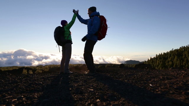 Hiking people reaching summit top giving high five at mountain top at sunset. Happy hiker couple.