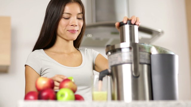 Woman making apple juice on juicer and drinking it fresh at home in kitchen. Juicing and healthy eating happy woman making green vegetable and fruit juice. Mixed race Asian model looking at camera.