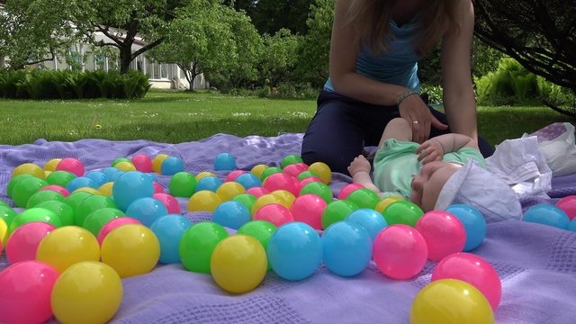 Mother dress up baby daughter cloth between colorful balls on plaid lay on meadow grass. Right side sliding shot. 4K
