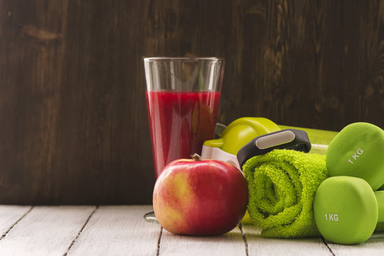 Fitness Or Diet Concept: Dumbbells, Fresh Red Smoothie, Apple, Towel And Activity Tracker Over Wooden Background. Toned Image. Selective Focus