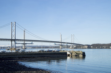 The South Queensferry end of the Forth Road Bridge, with two partially-completed towers and deck of the new Queensferry Crossing Bridge. South Queensferry Harbour is the foreground.