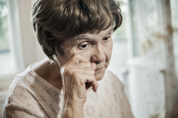 an elderly woman sits at the window and wipes away a tear