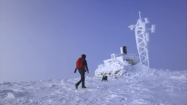 A hiker and their dog approaching a frozen weather station on the summit of Cairn Gorm in the Scottish Highlands on a cold sunny winters day.