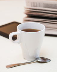 White cup of tea, digital tablet and spoon on light wooden desk.