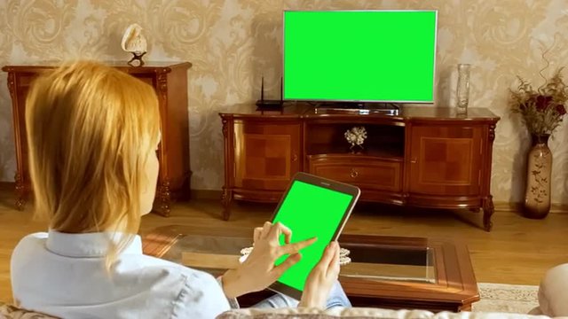 Young woman using tablet and watching television in living room. Shoot behind model's shoulders