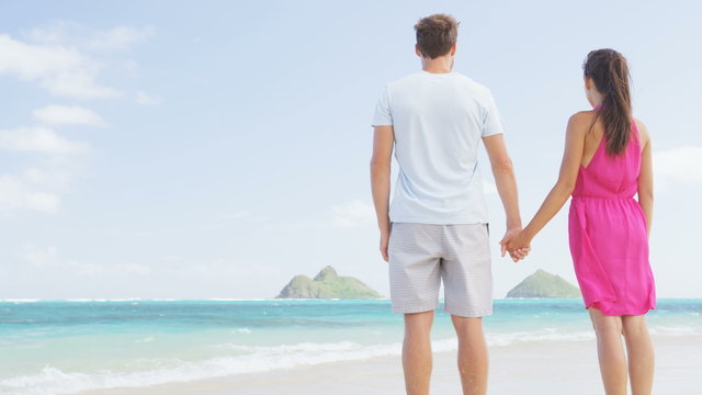 Beach vacation couple relaxing on summer holidays. Young people standing from behind holding hands looking at the ocean on Lanikai beach, Oahu, Hawaii, USA with Na Mokulua Islands. RED EPIC