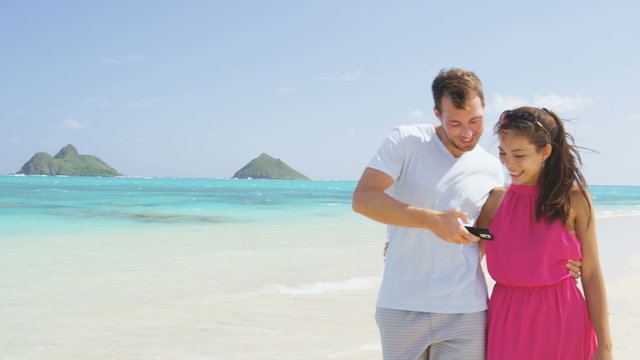 Man showing woman picture on phone app on beach using smartphone. Romantic couple in love on honeymoon having fun on outside on beach holding smart phone walking and laughing. 