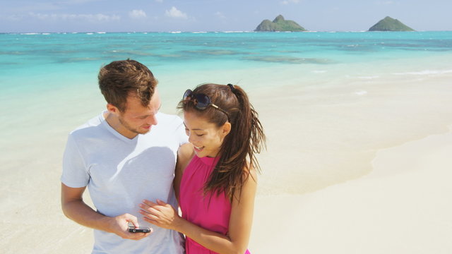 Couple on beach vacation using smart phone app. Romantic couple in love on honeymoon having fun on Lanikai beach, Oahu, Hawaii. Couple holding smartphone. RED EPIC SLOW MOTION.