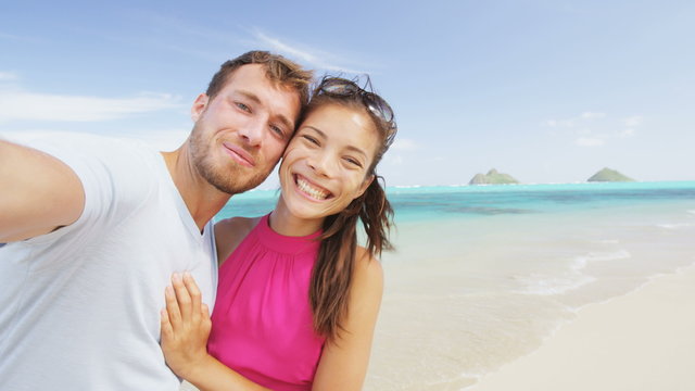 Couple relaxing on beach taking selfie picture with camera smart phone. Young multiracial couple on getaway vacation in Hawaii having fun looking at camera. Candid closeup. RED EPIC SLOW MOTION.