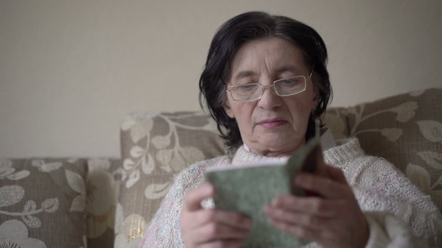 Old woman in glasses reading a prayer book on a sofa