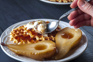 Waffles, pears cooked in sugar syrap and ice cream on white plate on blue wooden background. Woman hands with spoon