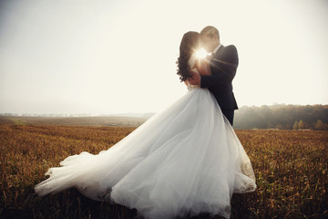 Romantic fairytale newlywed couple hug & kiss in field at sunset