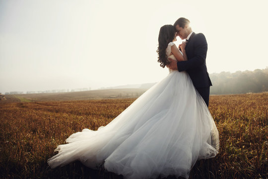 Romantic fairytale newlywed couple hug & kiss in field at sunset