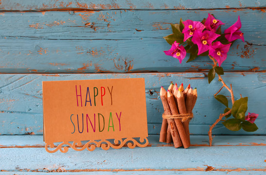vintage card with phrase: happy sunday and stack of wooden colorful pencils on wooden texture table next to purple bougainvillea flower.

