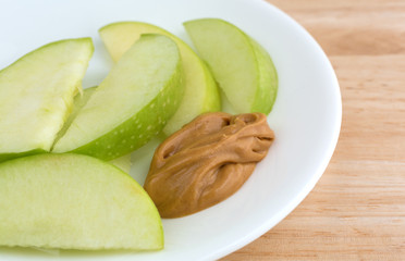 Side close view of green apple slices on white dish with peanut butter for dipping on a wood table top