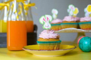 Colorful cupcake decorated with bunny picker on the yellow plate and orange juice in glasses in Easter scene 