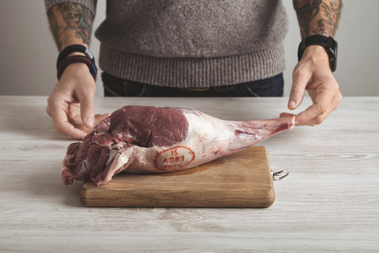 Tattooed butcher holds raw lamb leg in hands