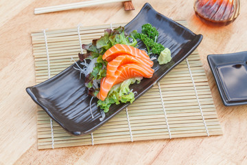 Sashimi salmon with vegetable and radish in black plate and chopsticks on wooden table.