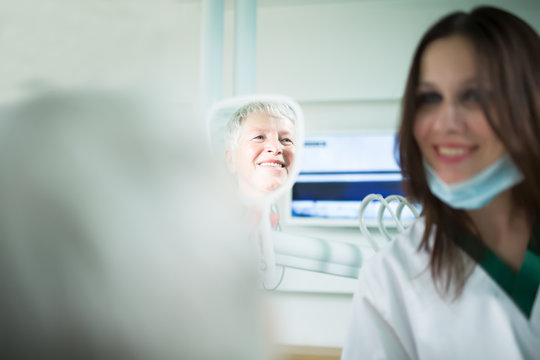 Old woman visiting the dentist taking care of her teeth.Young female dentist showing a granny her teeth,reflection in the mirror.Dentist doctor talking to a senior woman patient.Dental care for elder