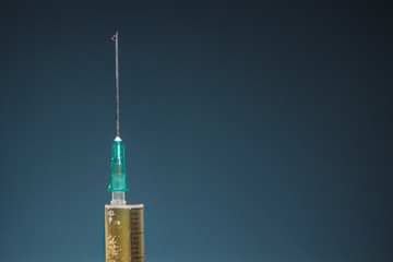 Hypodermic needle with a drop at its tip and the top of syringe with dark yellow drug, on dark blue...
