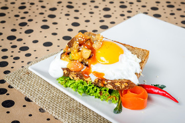 Fried egg with leaking yolk, mixed spicy vegetables on top of healthy toast, served on white plate with tomato, chili and green salad, copy space