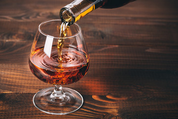 Pouring cognac or whiskey from the bottle into the glass against wooden background