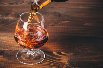 Pouring cognac or whiskey from the bottle into the glass against wooden background