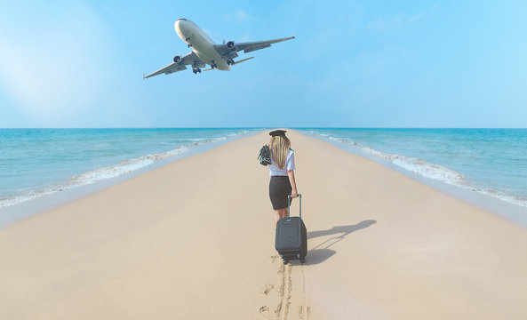 Travel concept. Young woman in flight attendant clothes walking on the beach with suitcase and hat . Overhead fly plane.