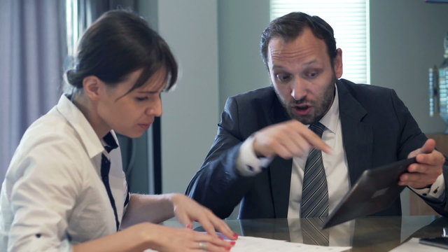 Boss with tablet yelling at businesswoman by the table in the office
