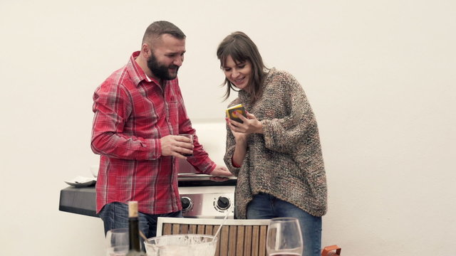 Two happy friends standing with smartphone during grill at terrace
