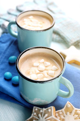 Two mugs of hot cacao with marshmallow, cookies and napkin on blue table