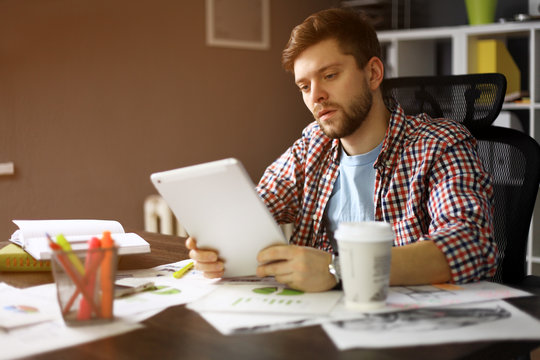 Thoughtful Male Person Looking To The Digital Tablet Screen While Sitting In Modern Interior At The Table, Experienced Entrepreneur Reading Some Text Or Electronic Book At The Office