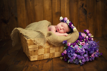 Newborn baby girl with a wreath in a wicker basket with a bouquet of purple wild flowers