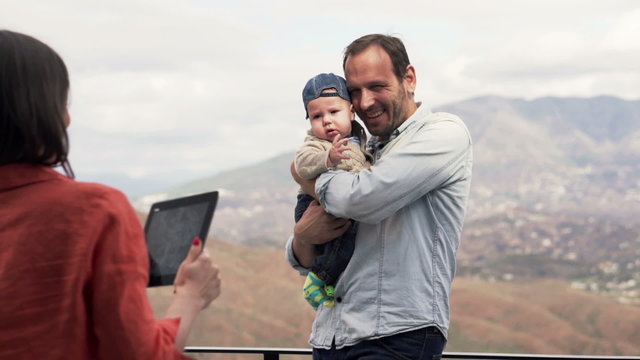 Young mother taking  photo with tablet of her small son and husband standing on terrace with splendid view
