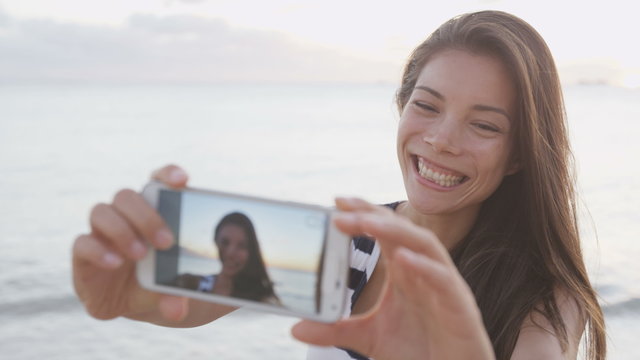 Girl taking selfie of herself using smartphone having fun outside on beach. Smart phone screen showing as woman takes selfies self portrait photos outdoors at beach sunset. RED EPIC SLOW MOTION.