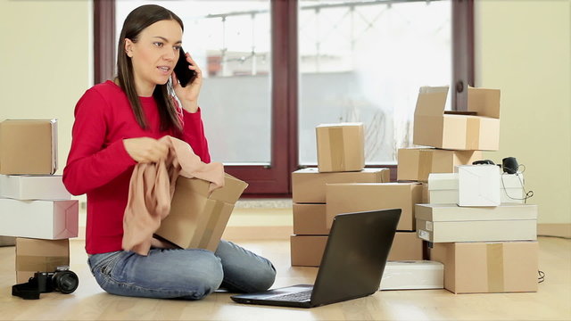 Young woman looks disappointed after shopping on line and complaining by phone with laptop and stack of boxes on the floor