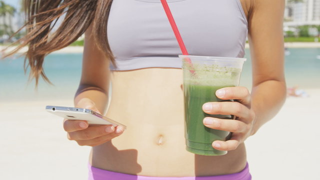 Smart phone Woman drinking vegetable Green detox smoothie using smartphone after fitness running workout on summer day. Fitness and healthy lifestyle concept with beautiful fit Asian Caucasian model.
