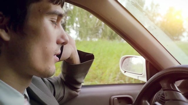 Young handsome dark-haired man driving car in the field and speaking on mobile phone in slowmotion. HD. 1920x1080