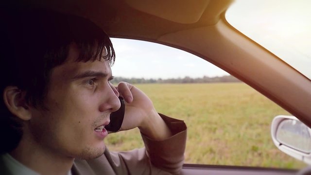 Young handsome dark-haired man driving car in the field and speaking on mobile phone in slowmotion. HD. 1920x1080