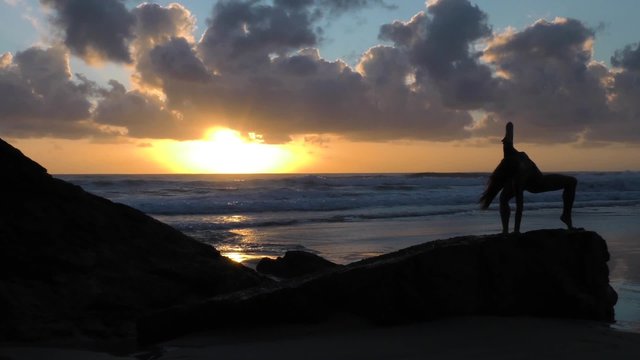 Attractive woman doing pilates on beach rock at sunrise. Slide.