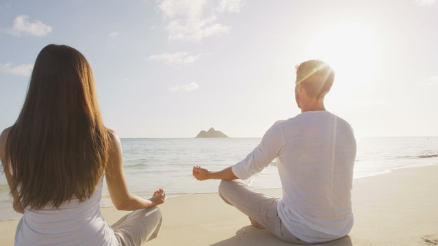 Yoga couple meditating in lotus pose relaxing outside on beach at sunrise. People woman and man in meditation in serene ocean landscape. Lanikai beach, Oahu, Hawaii, USA.
