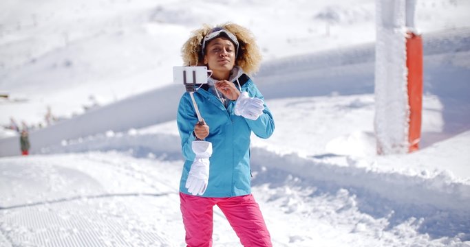 Smiling young woman posing for a photograph waving at her mobile phone camera as she takes a selfie on a stick in thick white snow